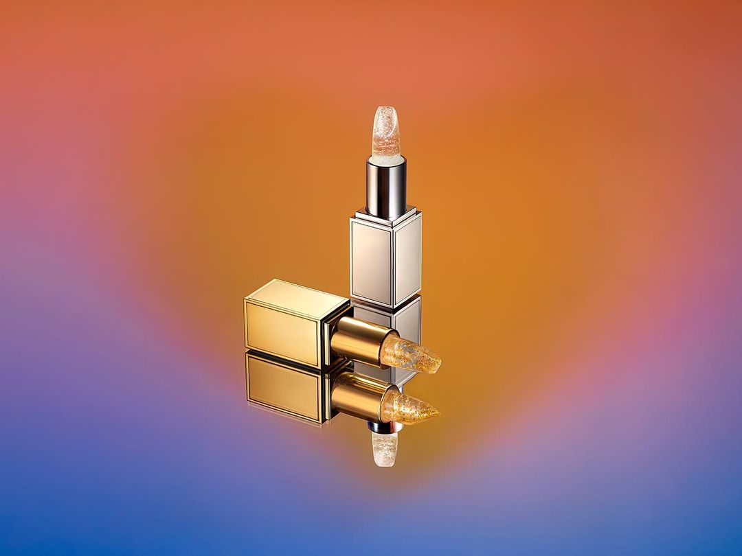 Two gold and silver metallic lipstick tubes on a reflective surface against a gradient background.