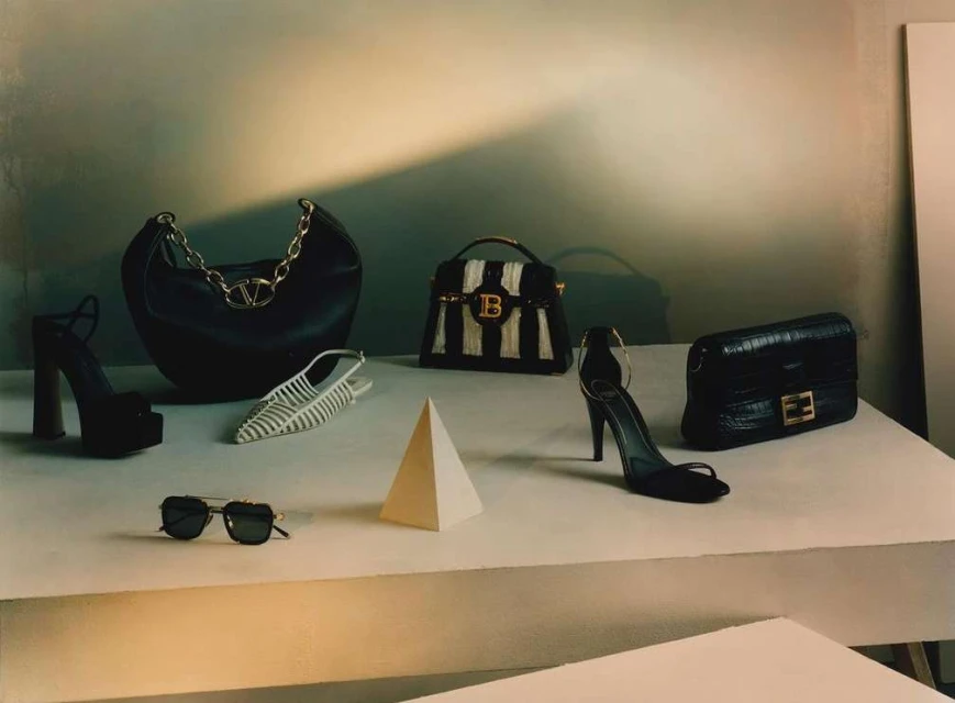 Fashion still life: Black leather handbags and high heel shoes from designer brands arranged on a stepped platform with a pyramid prop.