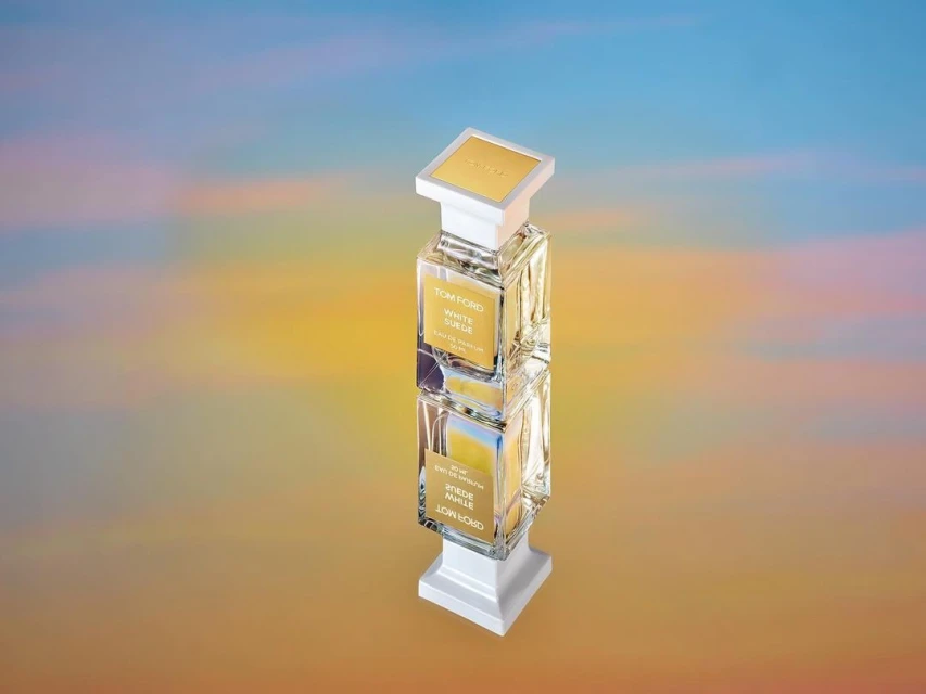 Tom Ford White Suede fragrance bottle on a mirrored surface with a diffused yellow and blue background, product shot, beauty.