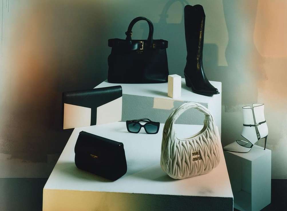 Fashion product laydown featuring an arrangement of handbags, boots, shoes, and sunglasses on a white multi-tiered platform.