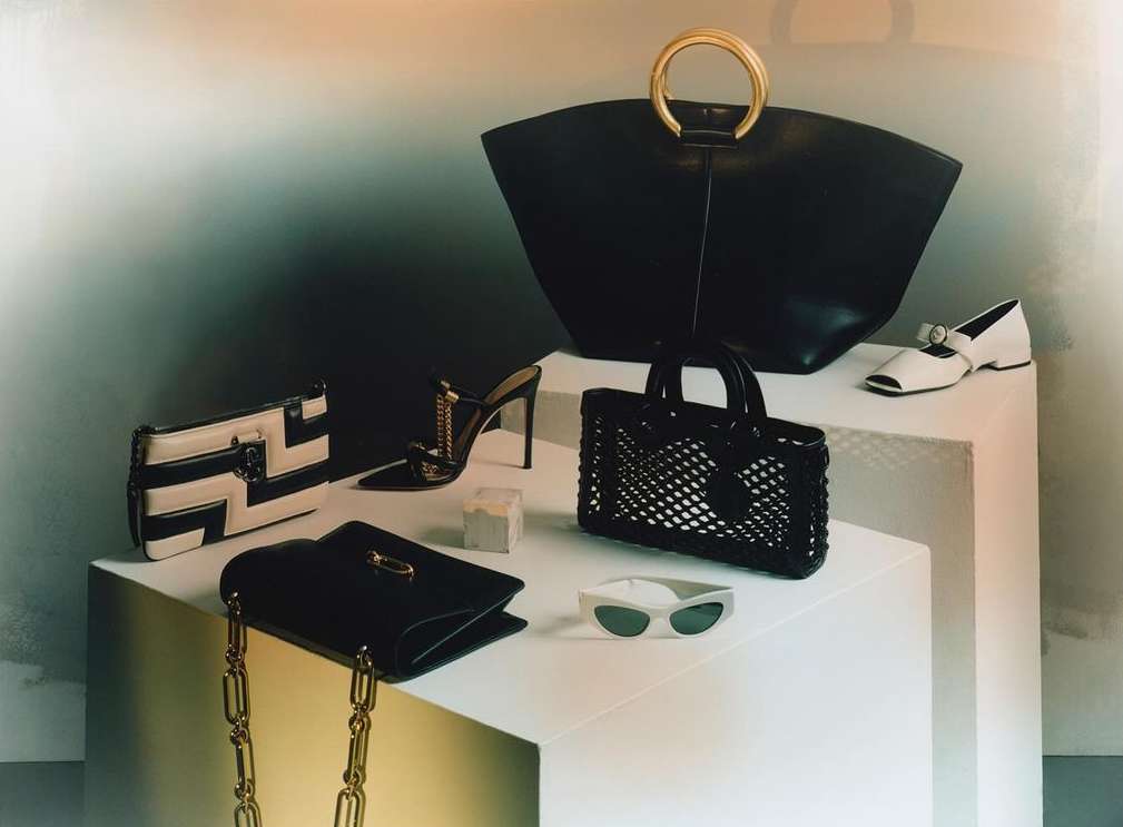 Fashion still life on stepped platform featuring multiple luxury designer handbags, pointed mule, Mary Jane, and cat-eye sunglasses.