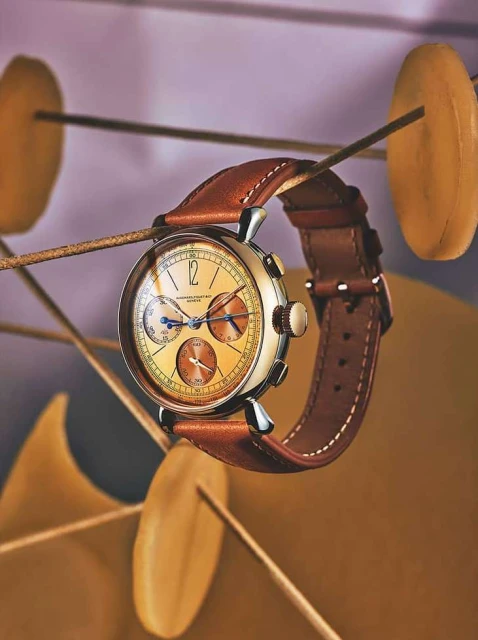 Close-up studio shot of an elegant wristwatch with a leather band, floating among abstract golden decorative shapes on a lavender background.