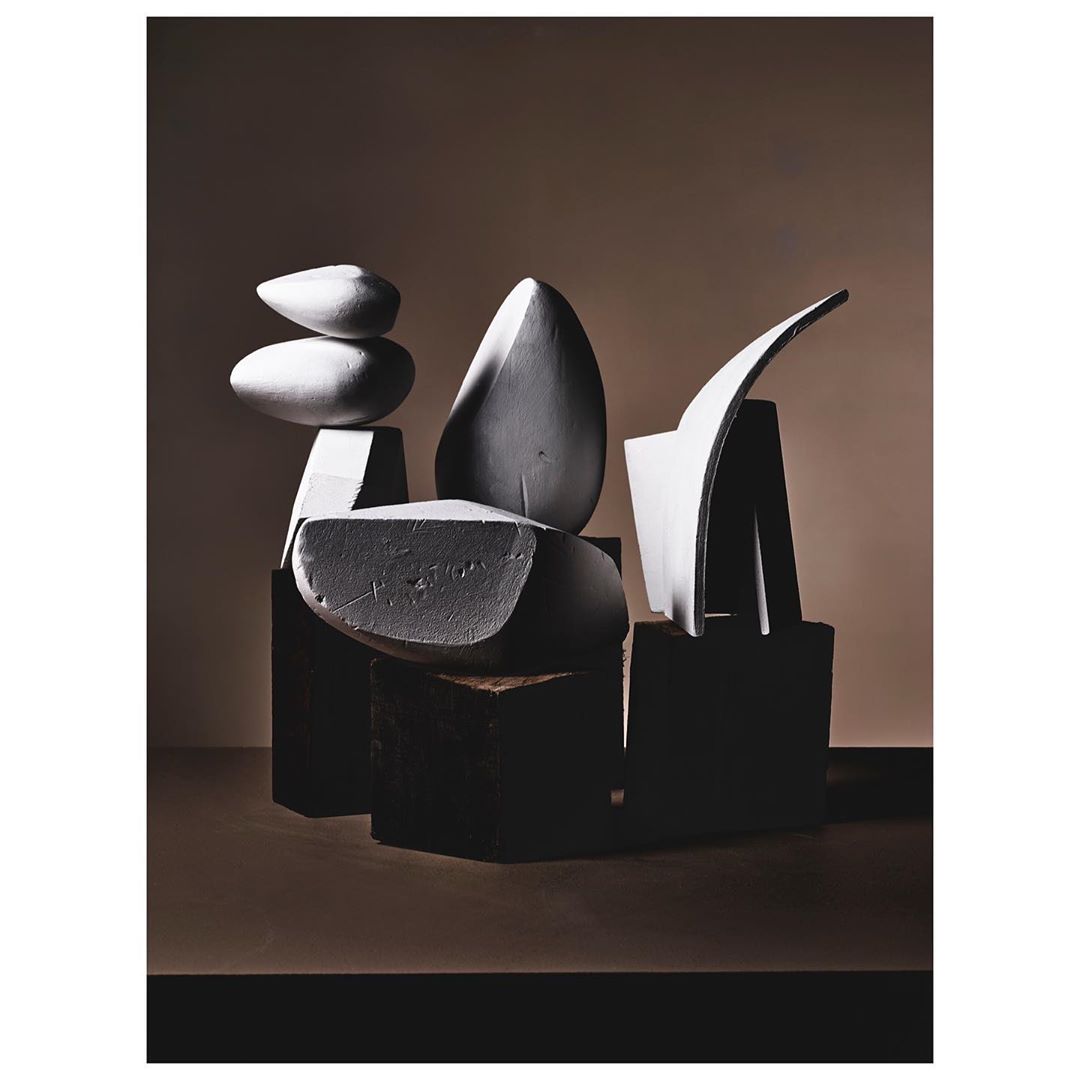 Abstract sculpture pieces in shades of white and black are arranged on a neutral tan background in a still life composition.