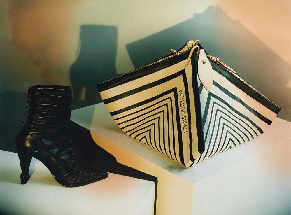 A fashion shot featuring a black leather ankle boot with a sculptural heel next to a Louis Vuitton striped handbag on a white surface.