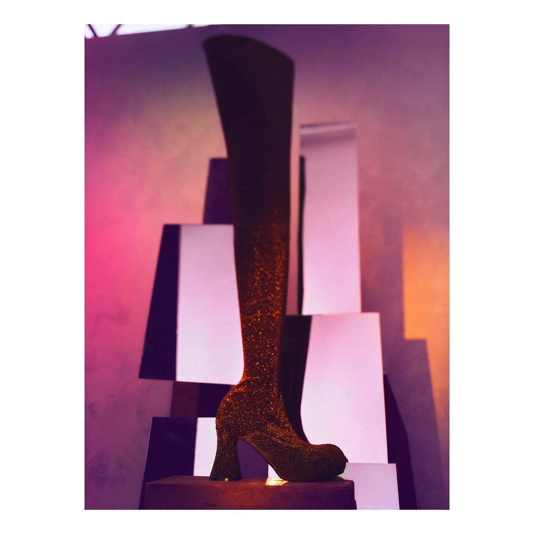 Glittery thigh-high boot on a geometric display with pink and purple lighting. Fashionable footwear in a studio setting.