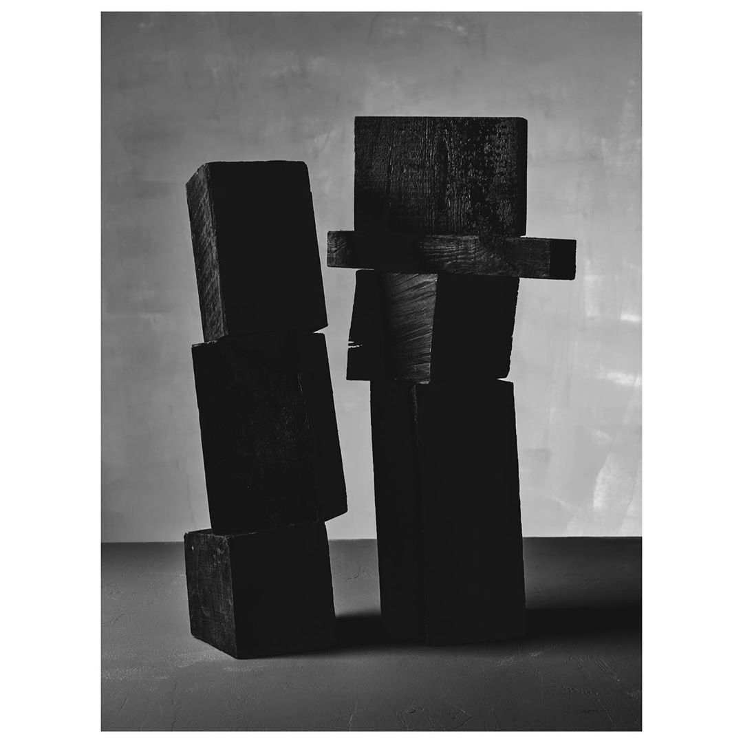 Monochrome photograph of stacked wood blocks against a mottled grey backdrop, arranged as an abstract sculptural form in a studio setting.