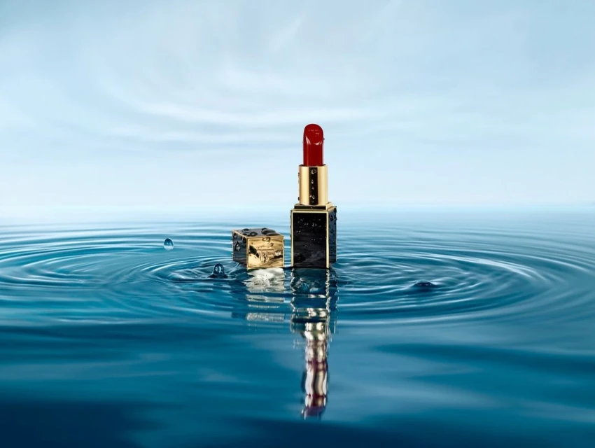 High-angle studio shot of red lipstick with gold accents standing in rippled blue water against a light-blue gradient sky backdrop.