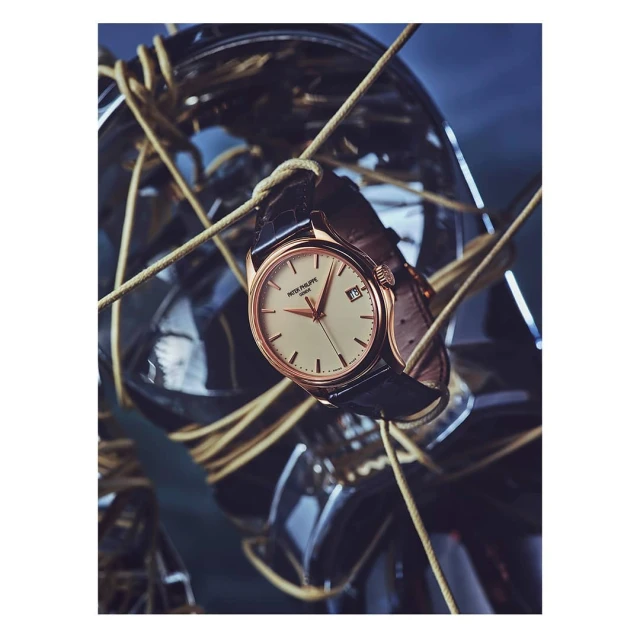 Wristwatch featuring a black leather strap and a cream-colored dial, displayed with tan cords and a metallic sphere in the background.