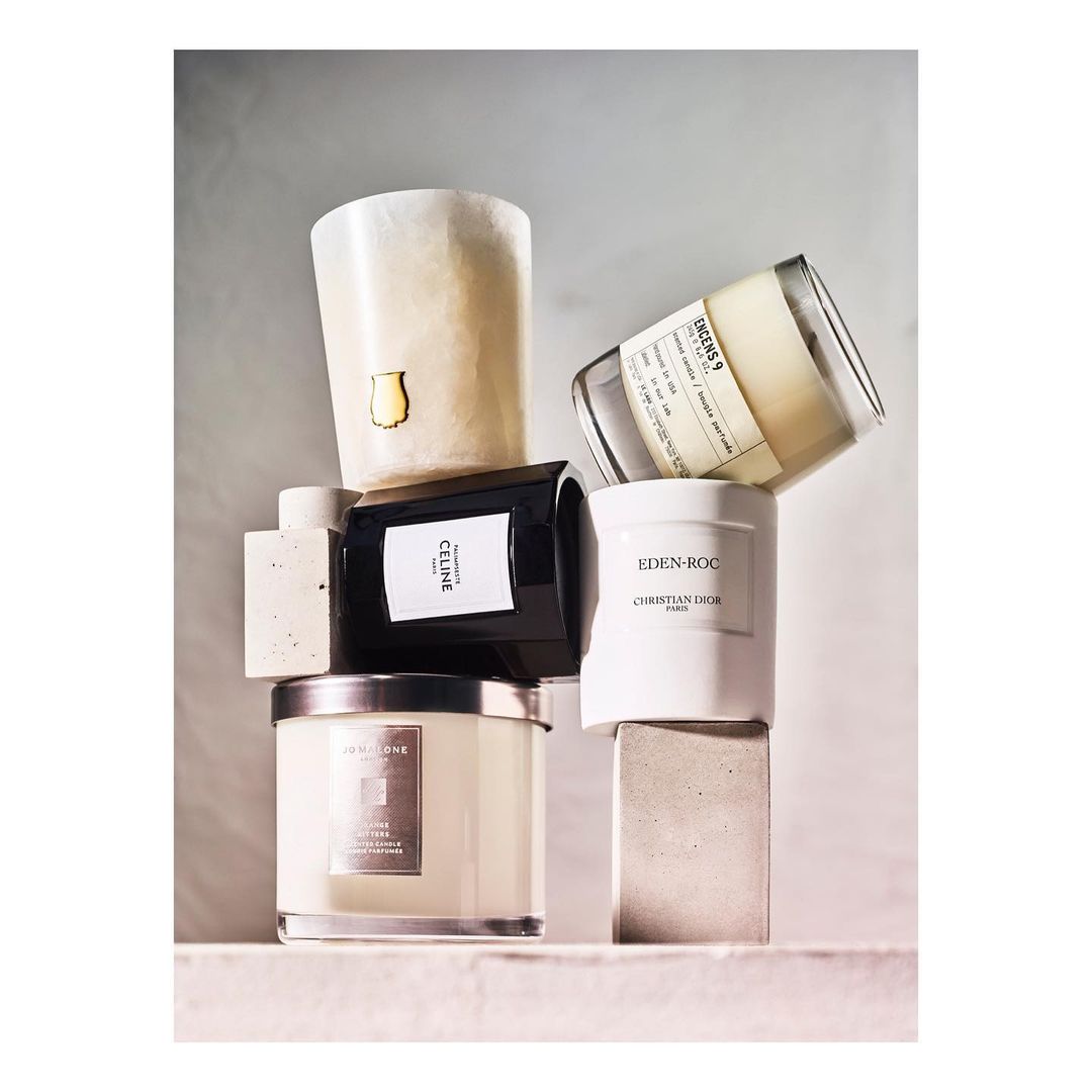 Assorted luxury candles from Celine, Christian Dior, and Jo Malone stacked on neutral concrete blocks in soft, diffused lighting.
