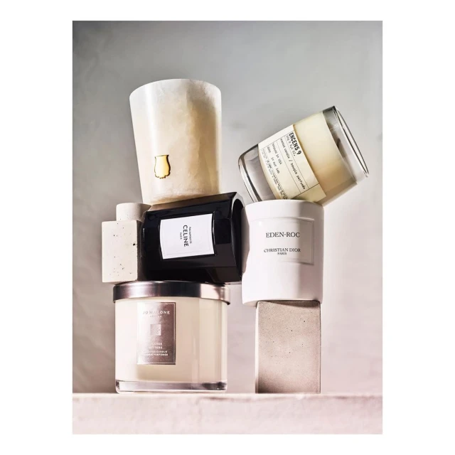 Assorted luxury candles from Celine, Christian Dior, and Jo Malone stacked on neutral concrete blocks in soft, diffused lighting.