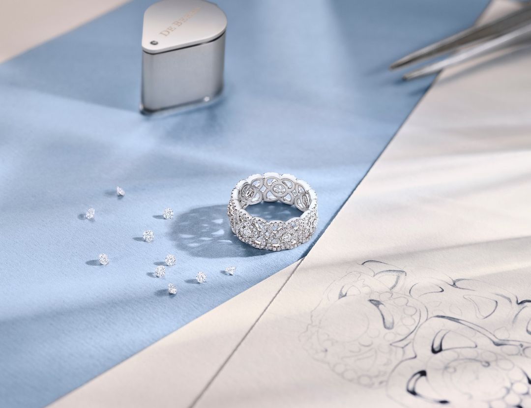 Close-up of a diamond ring surrounded by loose diamonds, a loupe, design sketches, and tweezers against a light blue backdrop.