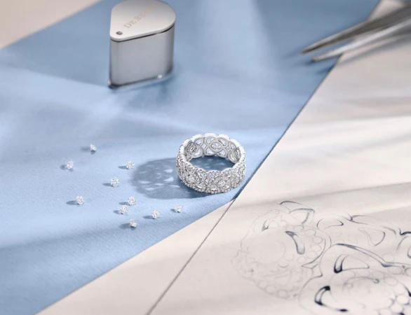 Close-up of a diamond ring surrounded by loose diamonds, a loupe, design sketches, and tweezers against a light blue backdrop.