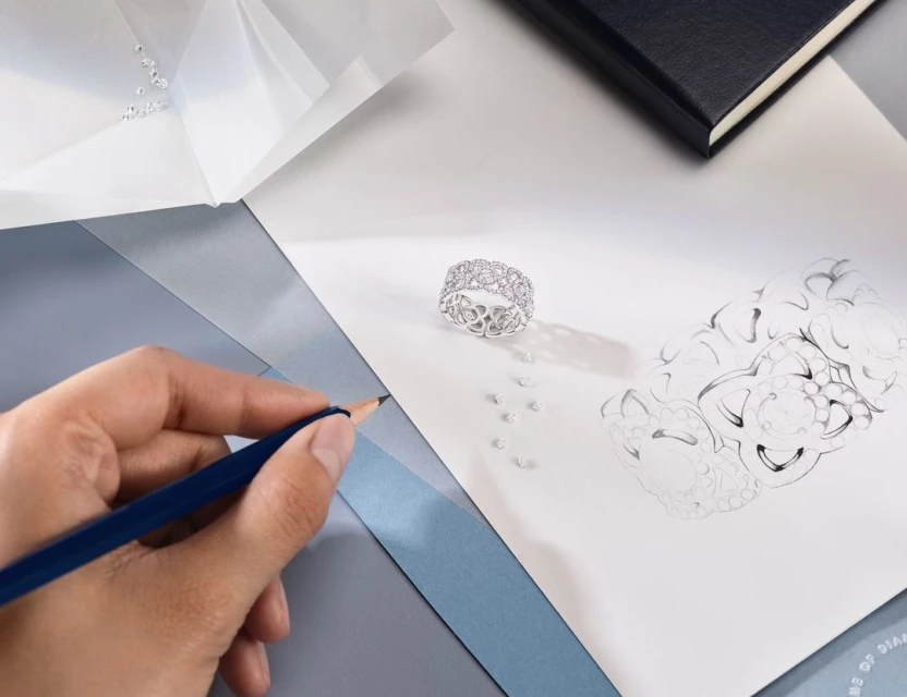 Close-up of a hand drawing a silver ring with diamonds on white paper, with loose diamonds and a black notebook in the background.