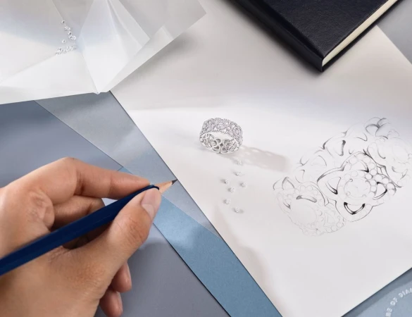 Close-up of a hand drawing a silver ring with diamonds on white paper, with loose diamonds and a black notebook in the background.
