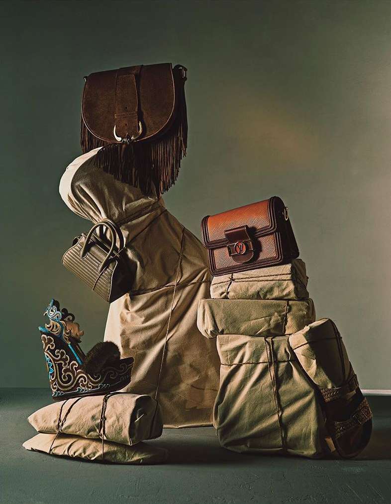 A collection of fashion handbags, totes, and shoes arranged in a stack with wrapped parcels, set against a muted green studio backdrop.