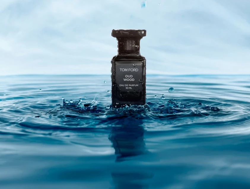 Tom Ford Oud Wood fragrance bottle sits in rippling blue water, with a blurred light-blue sky background and splashes from above.