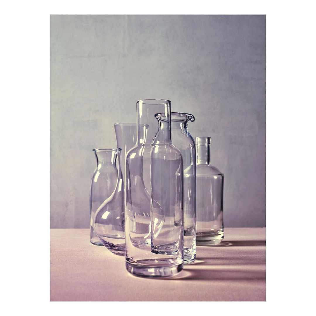 Still life studio shot of clear glass bottles and vases on a pink surface with a neutral textured background, minimalist aesthetic.