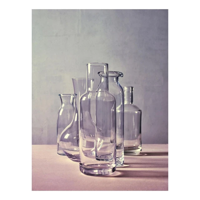 Still life studio shot of clear glass bottles and vases on a pink surface with a neutral textured background, minimalist aesthetic.