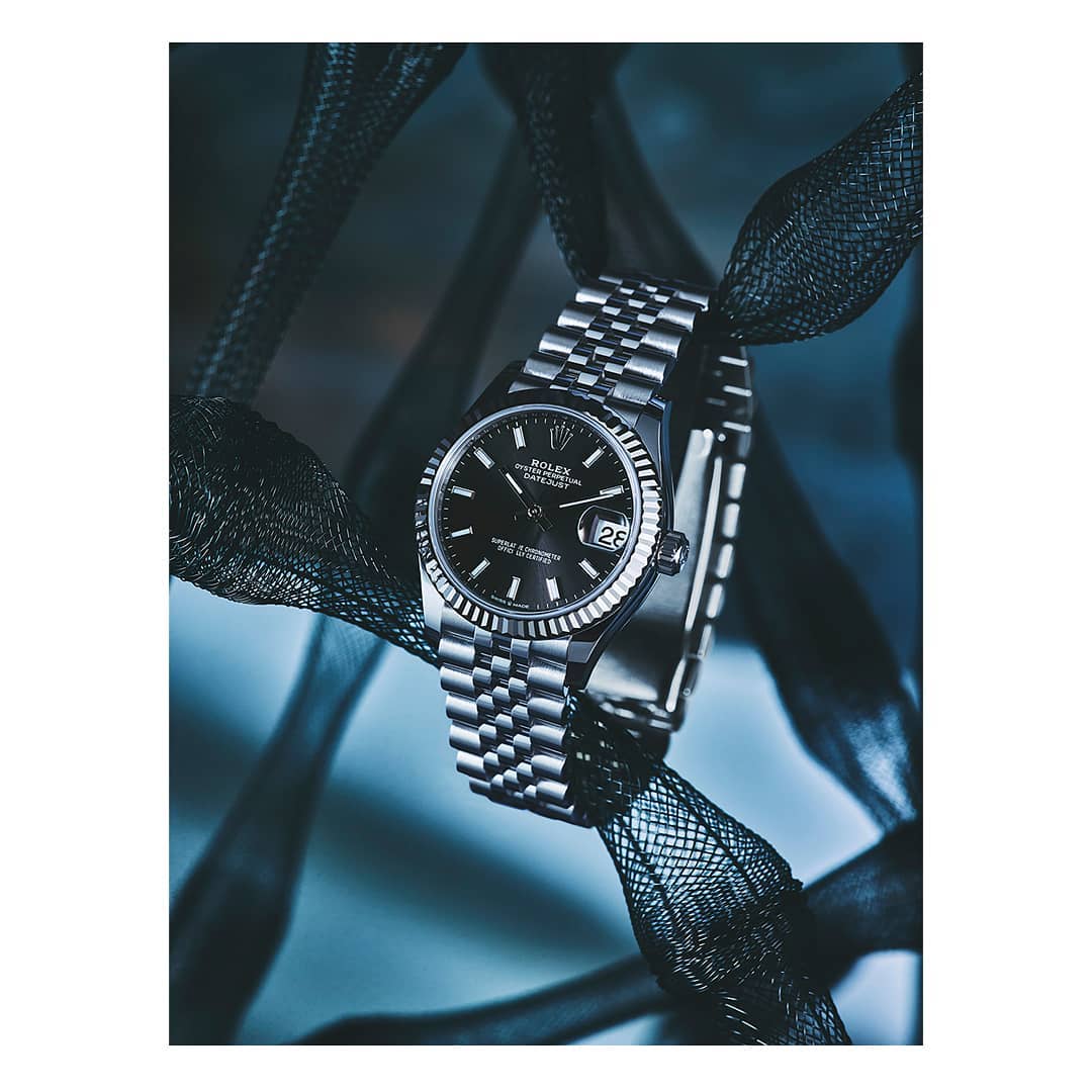 Close-up product shot of a Rolex Datejust wristwatch with a silver jubilee bracelet draped with black mesh fabric on a cool blue background.