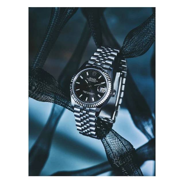 Close-up product shot of a Rolex Datejust wristwatch with a silver jubilee bracelet draped with black mesh fabric on a cool blue background.