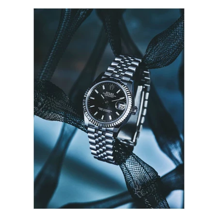 Close-up product shot of a Rolex Datejust wristwatch with a silver jubilee bracelet draped with black mesh fabric on a cool blue background.