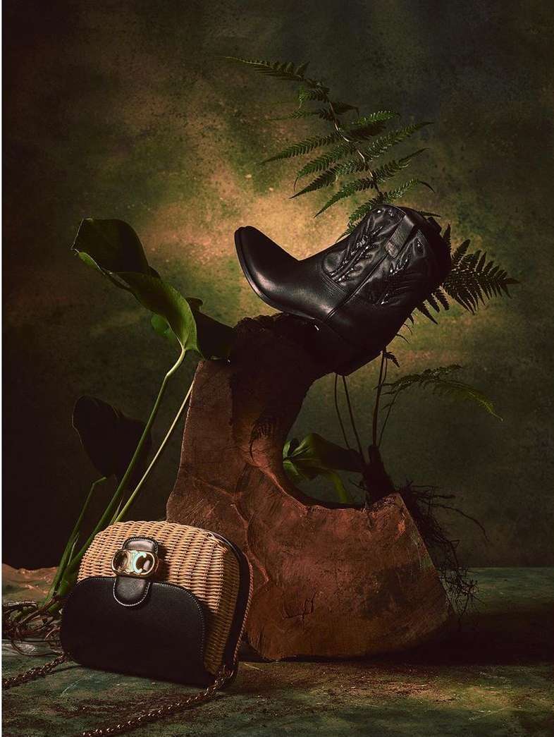 Still life photograph of a black cowboy boot, a straw purse with a black flap, and tropical plants against a textured green background.