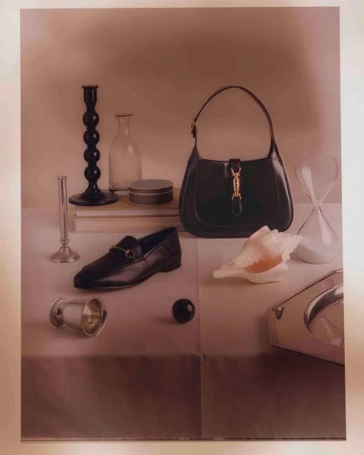 Still life with a black leather purse, a black leather loafer, an hourglass, and various decorative objects on a neutral surface.