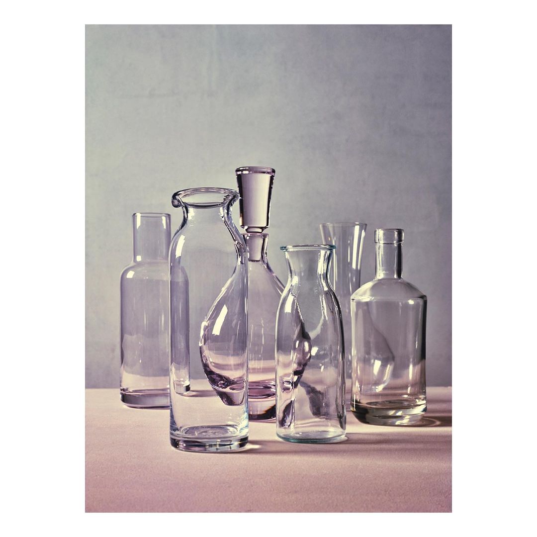Still life photograph featuring an arrangement of seven clear glass bottles in varying shapes and sizes on a light pink surface.