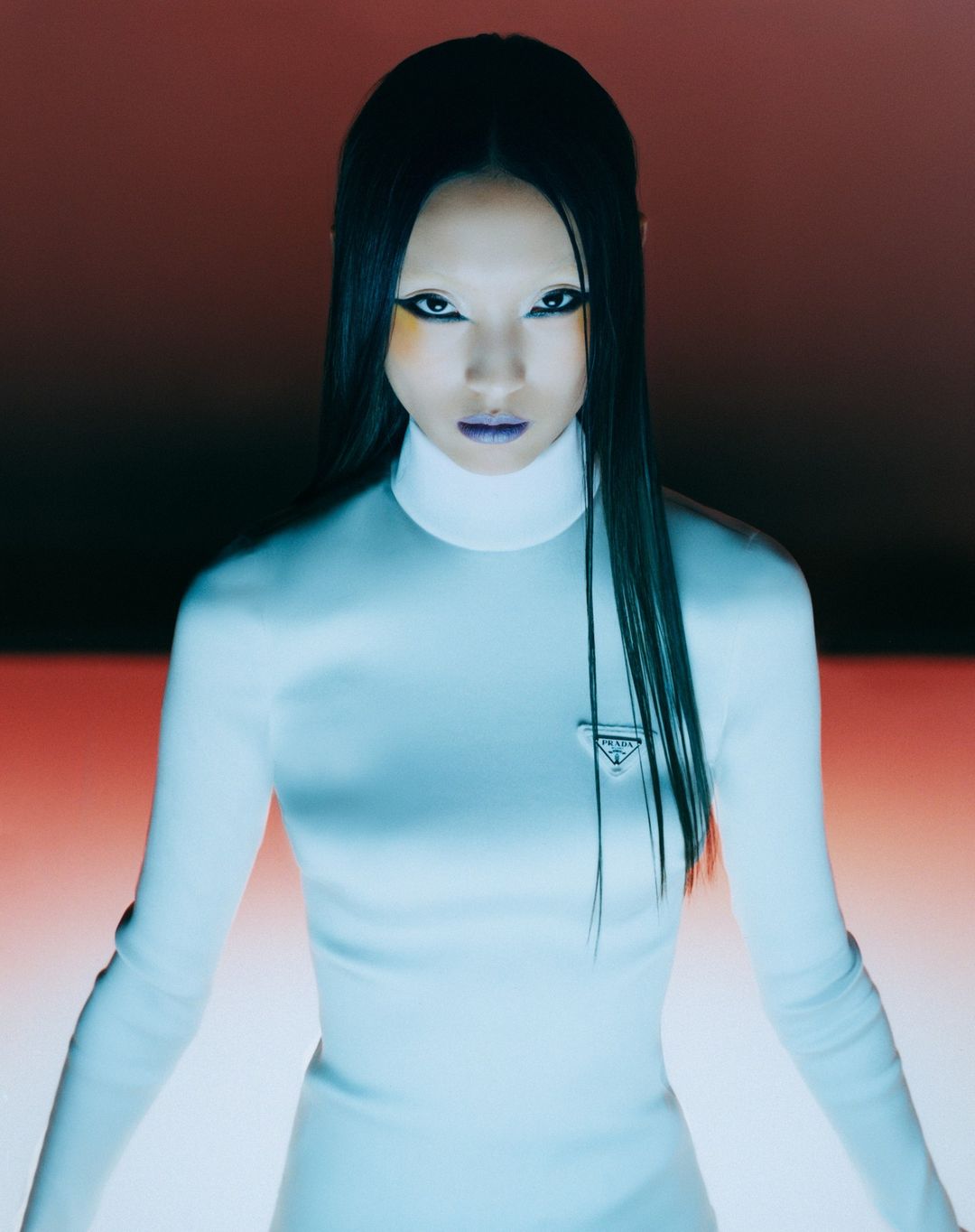 Woman with dark hair and fashion makeup in white turtleneck sweater with Prada logo, in a close-up studio shot with an orange background.