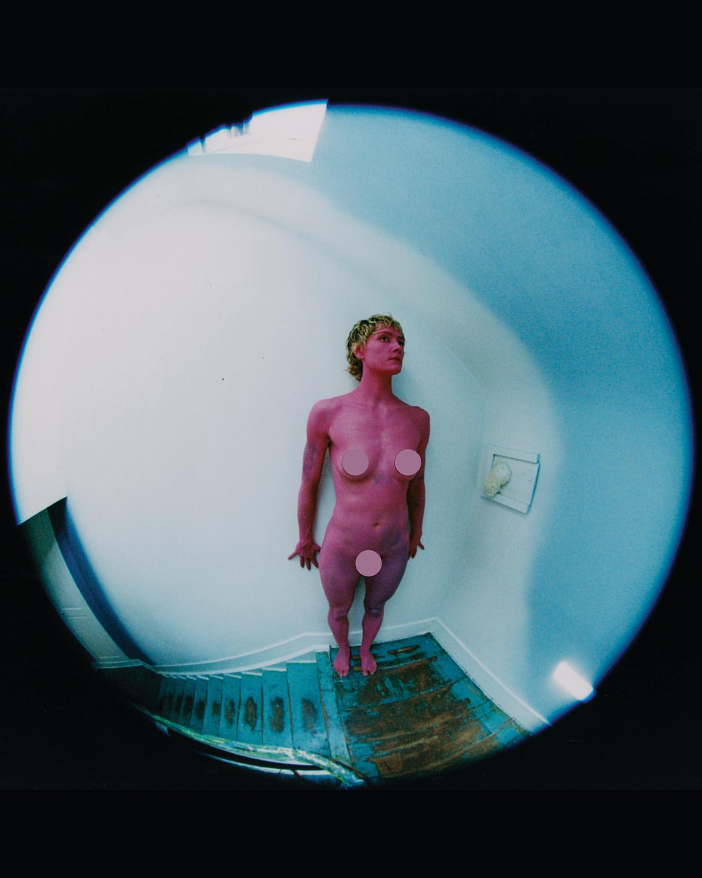 Nude adult female stands at the bottom of blue staircase in a fisheye indoor shot, pink body paint, blonde hair, and white wall. 