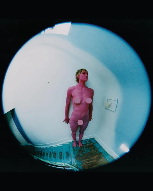 Nude adult female stands at the bottom of blue staircase in a fisheye indoor shot, pink body paint, blonde hair, and white wall. 