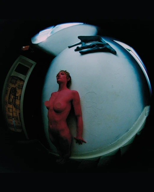 Full shot of a nude, pink-painted woman posing in front of a white wall, with a distorted fisheye lens effect in a dark indoor setting.