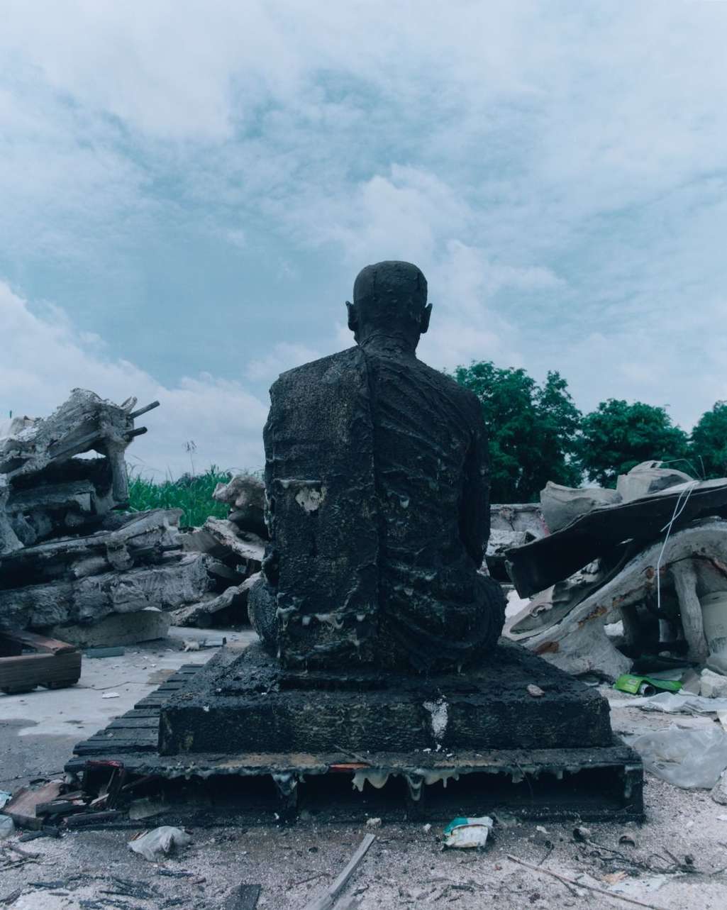Stone monk statue sits in outdoor debris-filled environment under overcast sky, creating somber mood with shades of gray and blue.