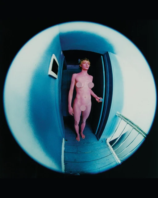 Full-length fisheye view of a naked woman in doorway with laurel wreath, shot indoors with blue walls and wooden floorboards.
