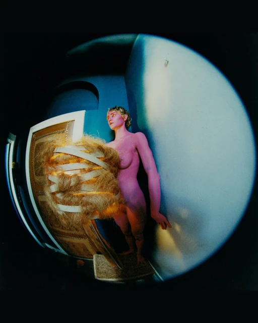 Woman strikes a pose next to a structure covered in tan straw and white tape in an indoor studio shot with a wide angle lens.