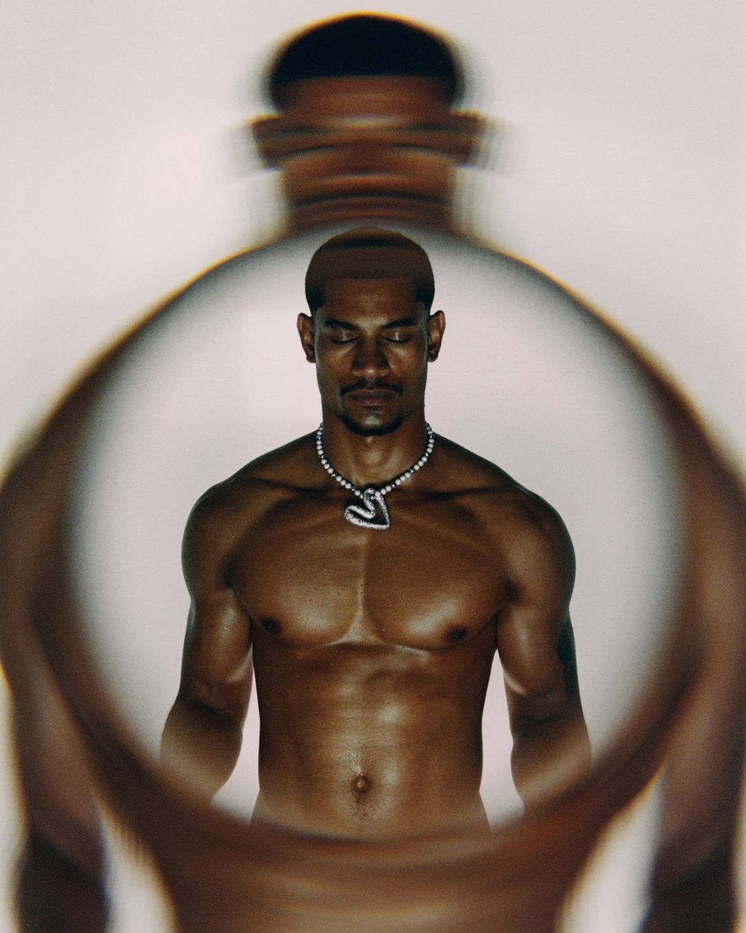 Portrait of adult male with closed eyes, necklace, muscular build, photographed through blurred glass in bright indoor lighting.