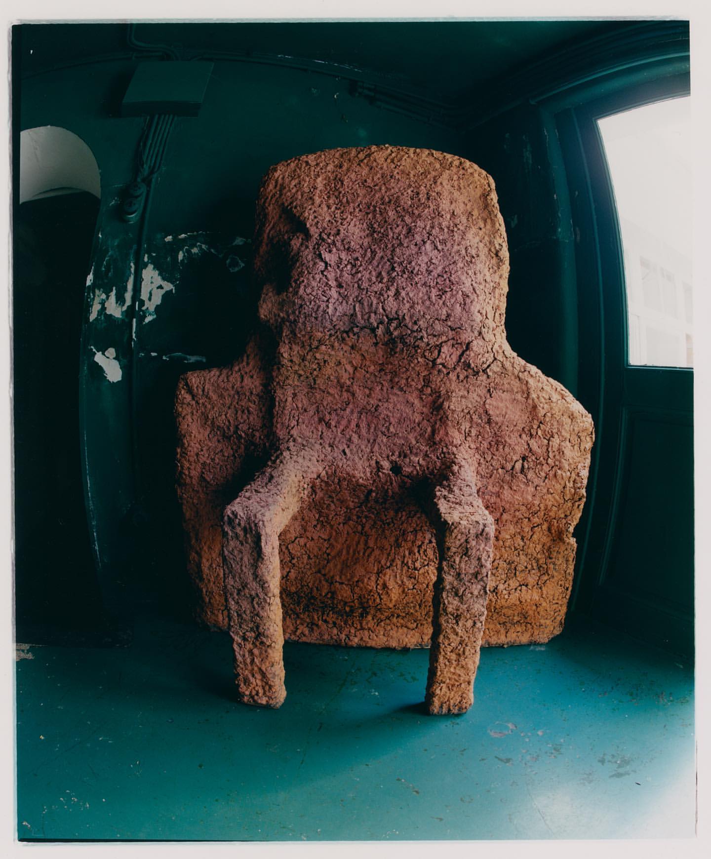 Abstract art photograph of a rough textured, vaguely humanoid sculpture, illuminated against a green interior with a distant window.
