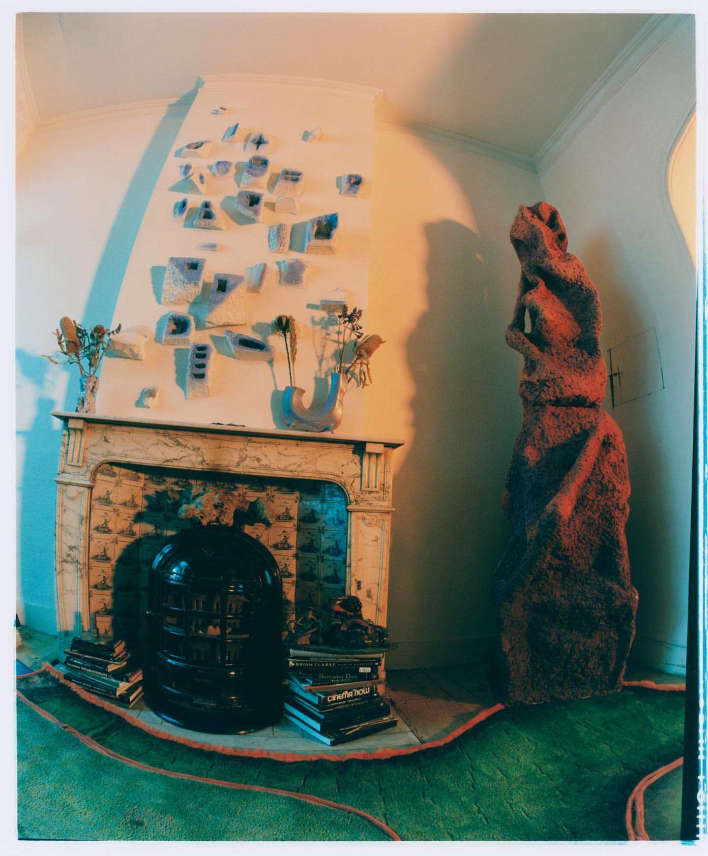 Eclectic living room featuring a fireplace with mantel art, an abstract sculpture, stacked books, and green flooring in soft lighting.