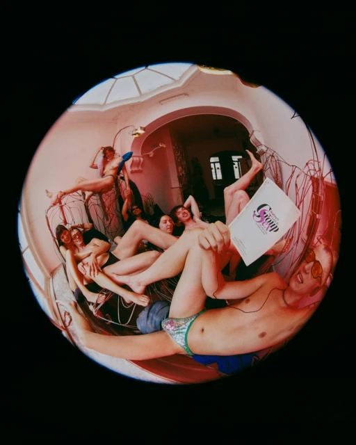 A group of shirtless adults lounge indoors in a circular fish-eye shot, one holding a document, bathed in natural light and red tones.