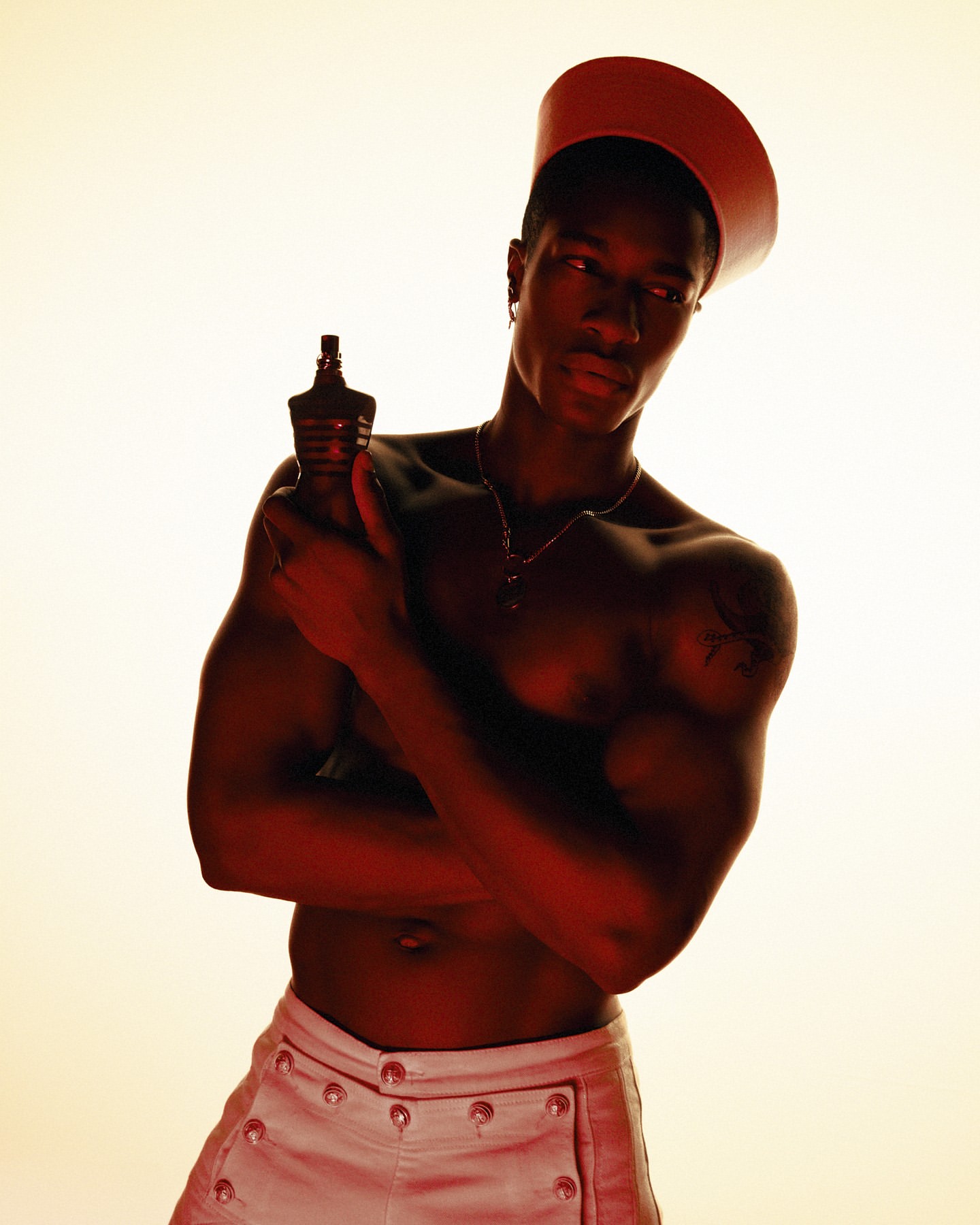 An adult male with dark skin, wearing a sailor cap and white pants, holds a bottle of cologne in a studio shot with warm lighting.