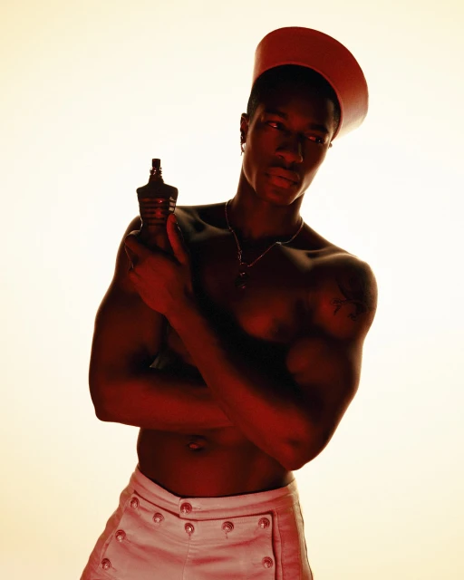 An adult male with dark skin, wearing a sailor cap and white pants, holds a bottle of cologne in a studio shot with warm lighting.