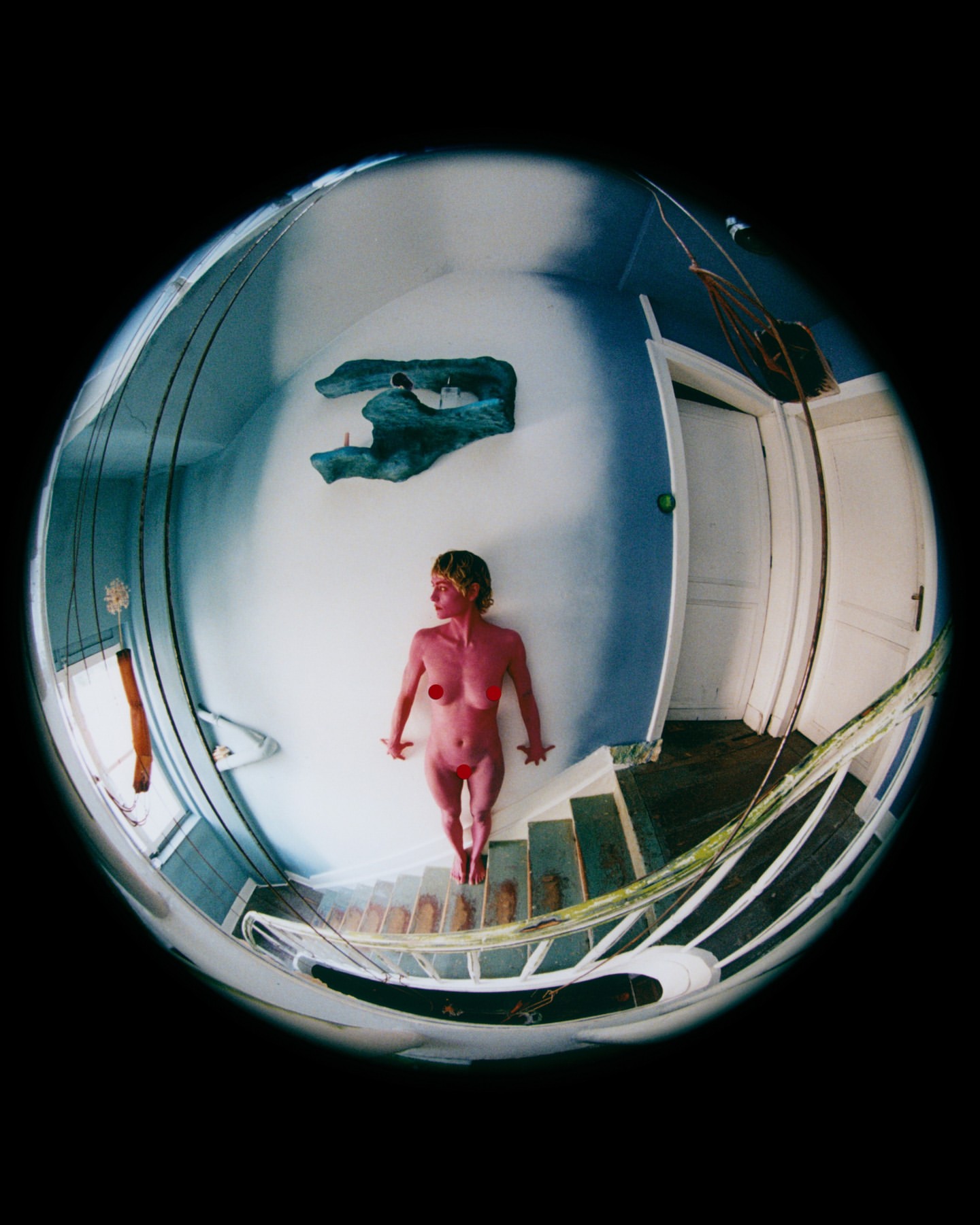 Nude adult woman standing on steps viewed through fisheye lens in indoor environment with a sculpture, blue walls and white door.