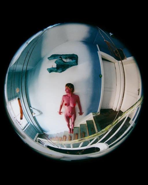 Nude adult woman standing on steps viewed through fisheye lens in indoor environment with a sculpture, blue walls and white door.