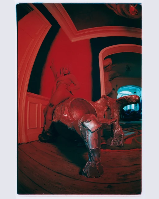 Photo of a woman on top of a sculpture resembling a horse inside a room with a red color cast and a wide angle lens.