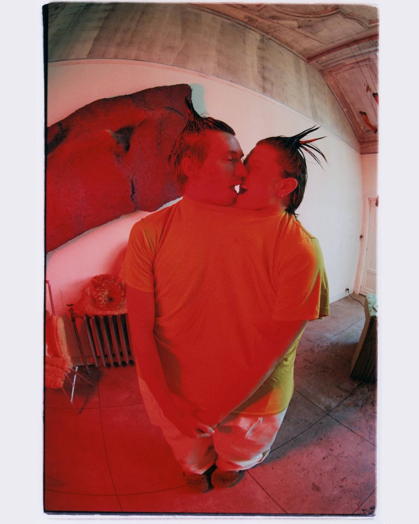 Two people kissing indoors, captured with a wide-angle lens distorting the room's architecture and featuring a red color cast over the scene.