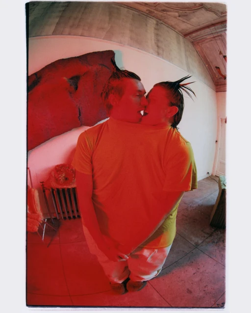 Two people kissing indoors, captured with a wide-angle lens distorting the room's architecture and featuring a red color cast over the scene.