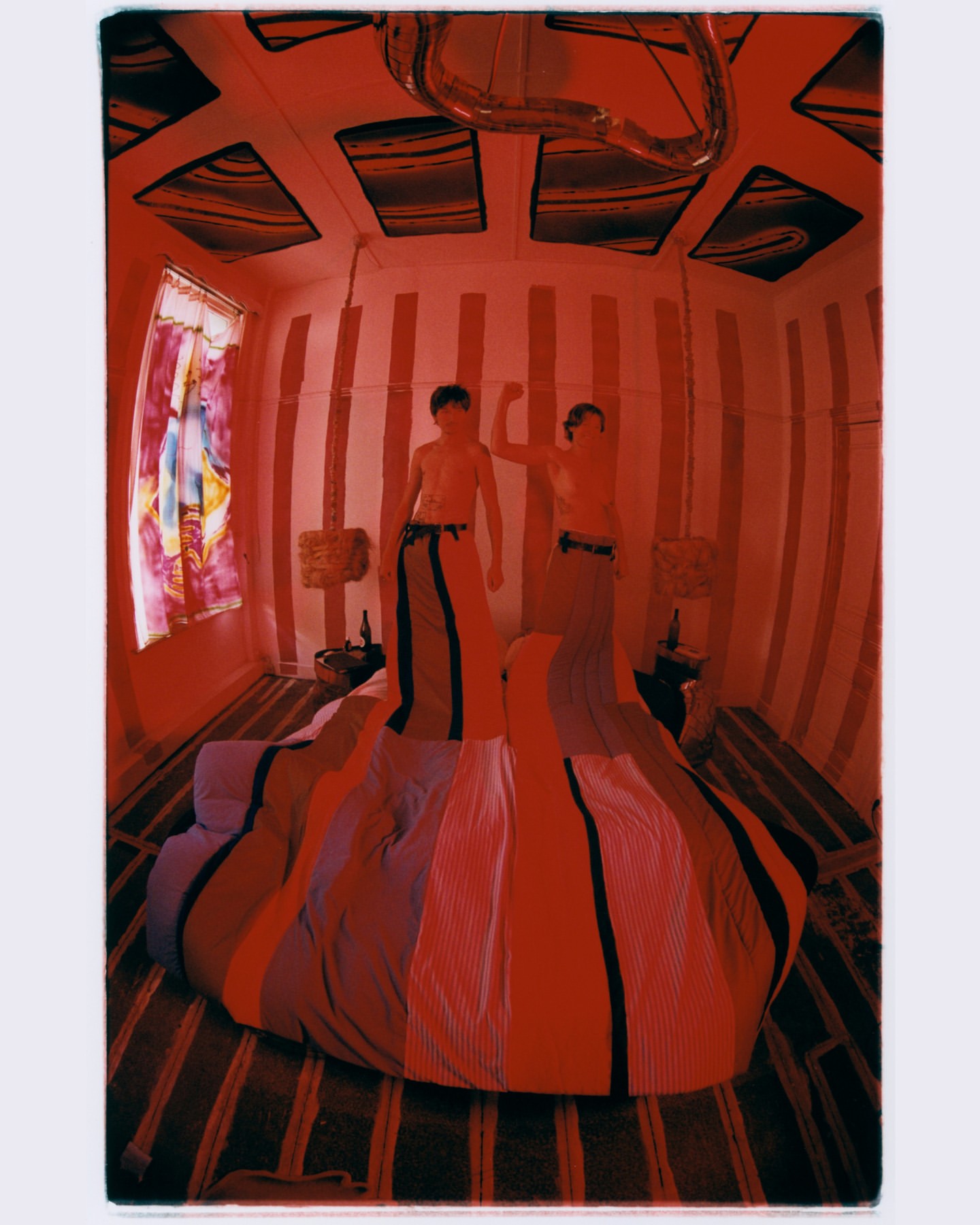 Indoor photo of two shirtless, young adult males wearing matching striped pants, standing on a striped bed in a vertically striped room.