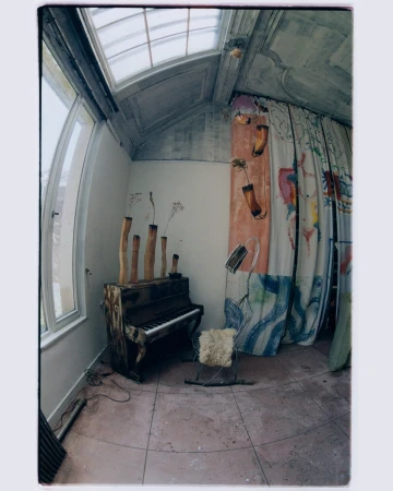 Photo of a weathered piano with wooden pipes as decor, a white chair, and abstract art in a sunlit, sparsely furnished room.