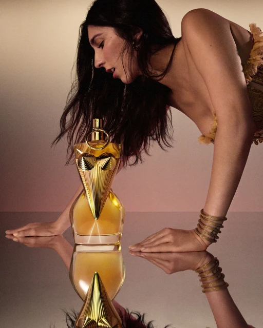 Brunette woman leans toward a golden perfume bottle with mirrored reflection in a beauty advertisement. Gold bracelets visible.