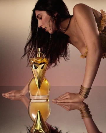 Brunette woman leans toward a golden perfume bottle with mirrored reflection in a beauty advertisement. Gold bracelets visible.